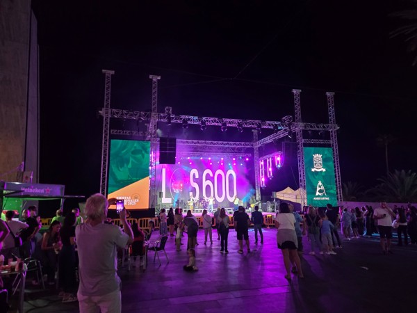 Escenario de un concierto nocturno en Adeje durante las fiestas patronales de 2025, con público delante y luces de colores iluminando la plaza.