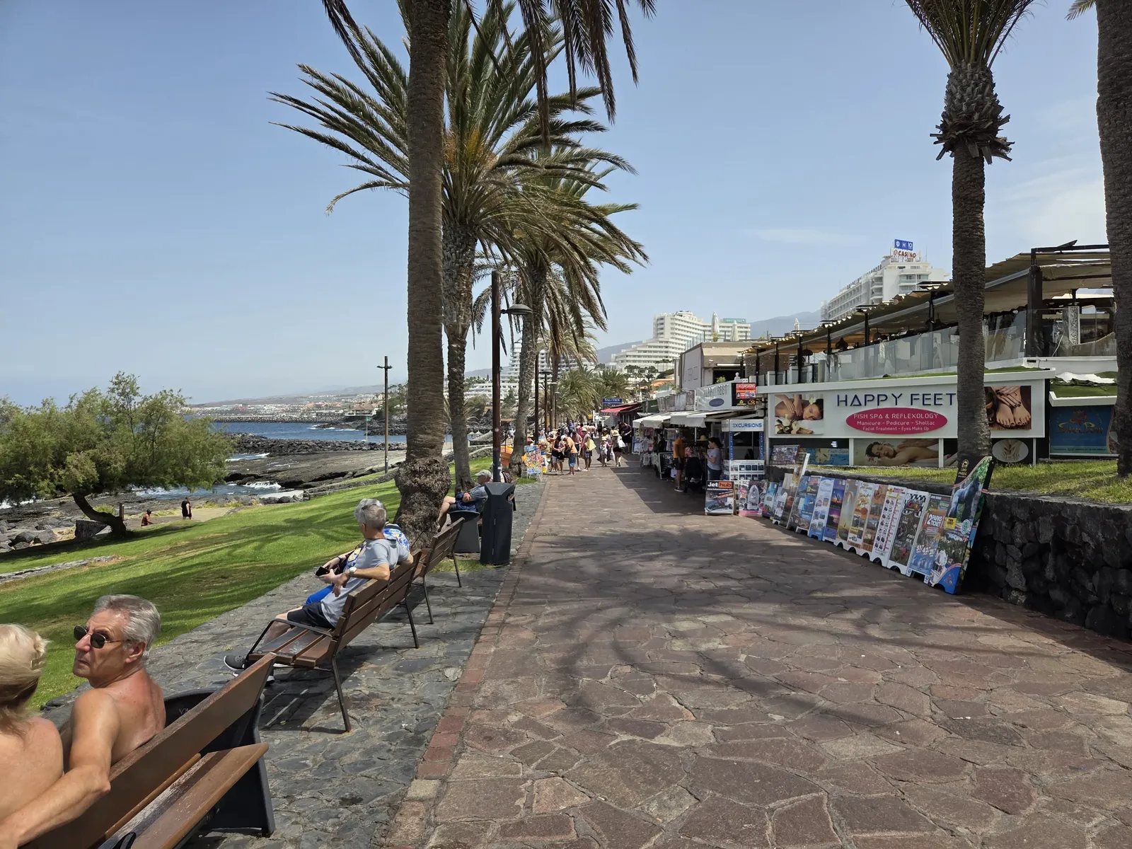 Paseo marítimo de Costa Adeje con palmeras, bancos y comercios abiertos hacia el océano