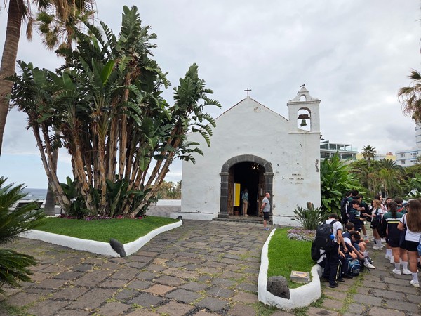 Pequeña ermita blanca junto al mar con palmeras y visitantes en un entorno tranquilo y tradicional.
