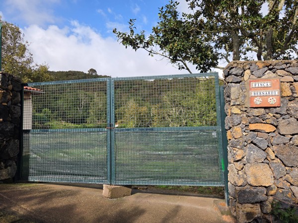 Entrada de una finca con gran portón metálico, muros de piedra volcánica y un cartel que dice Finca Bernardi, en un entorno rural y arbolado.