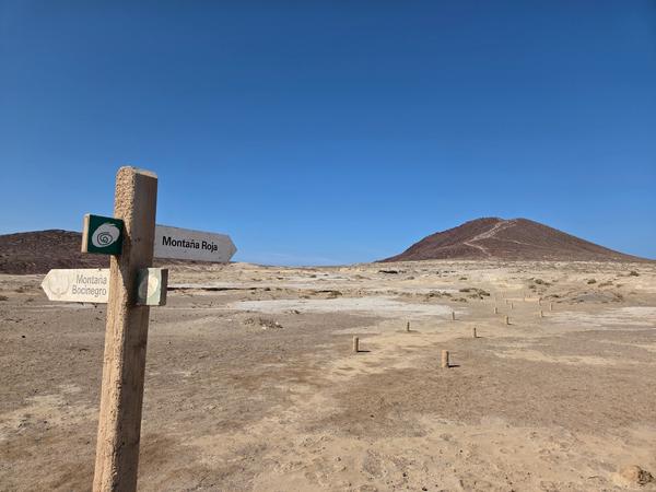 Señalización de senderismo indicando la dirección hacia Montaña Roja y Montaña Bocinegro, con el paisaje árido de fondo.