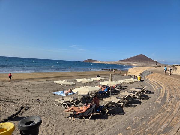 El Médano y Montaña Roja Playa de El Médano con sombrillas y hamacas, con la Montaña Roja al fondo y turistas caminando por la arena.