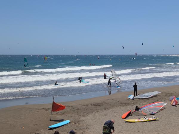 Windsurf en El Médano Personas practicando windsurf y kitesurf en la playa de El Médano con viento fuerte y olas.