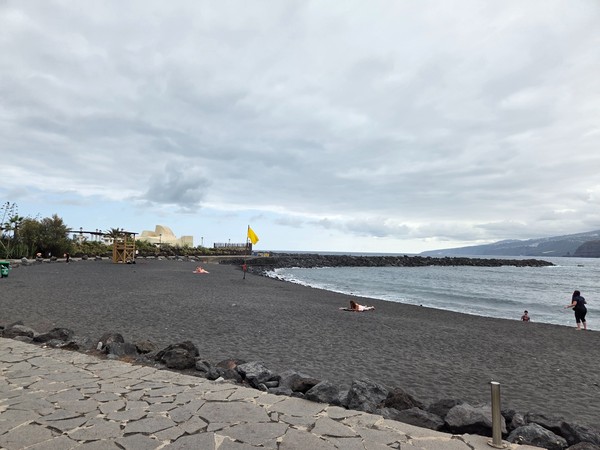 Playa Martiánez, Tenerife Playa de arena volcánica en Martiánez, Tenerife, con bandera amarilla, algunas personas tomando el sol y el auditorio de fondo.