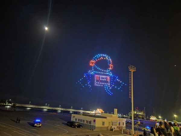 Espectáculo nocturno de drones en el cielo de Santa Cruz formando la figura de un astronauta, con la luna visible y el puerto al fondo.