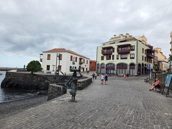 Zona costera de Puerto de la Cruz con esculturas, casas coloniales y turistas paseando bajo cielo nublado.