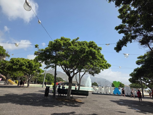 Plaza arbolada en Santa Cruz con fuente, personas paseando y el gran letrero decorativo de la ciudad.