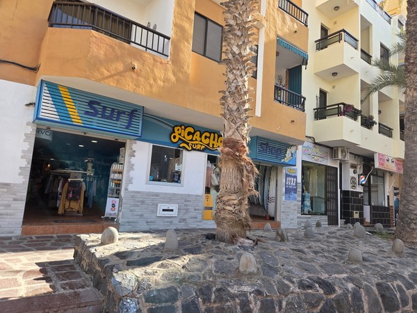 Tienda de surf en El Médano Tienda de surf en El Médano, Tenerife, con escaparate de ropa y tablas, en una calle peatonal con edificios de apartamentos.