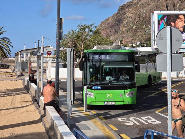 Autobús Titsa en parada junto a la playa Autobús verde de Titsa en la parada cerca de la playa, con personas descansando y paisaje montañoso de fondo.